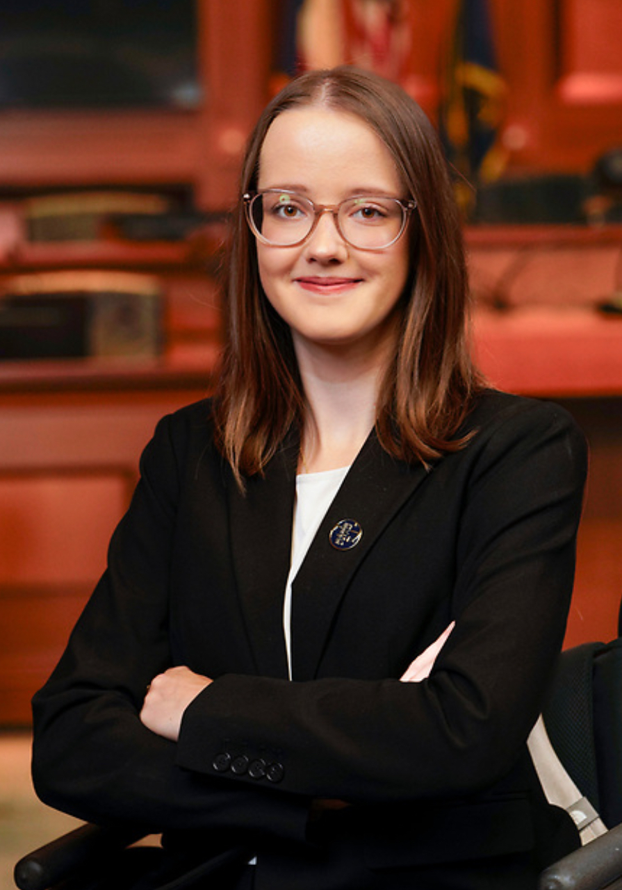 White woman with light brown hair and glasses, giving a soft smile. She sits with her arms crossed in front of a courtroom background.