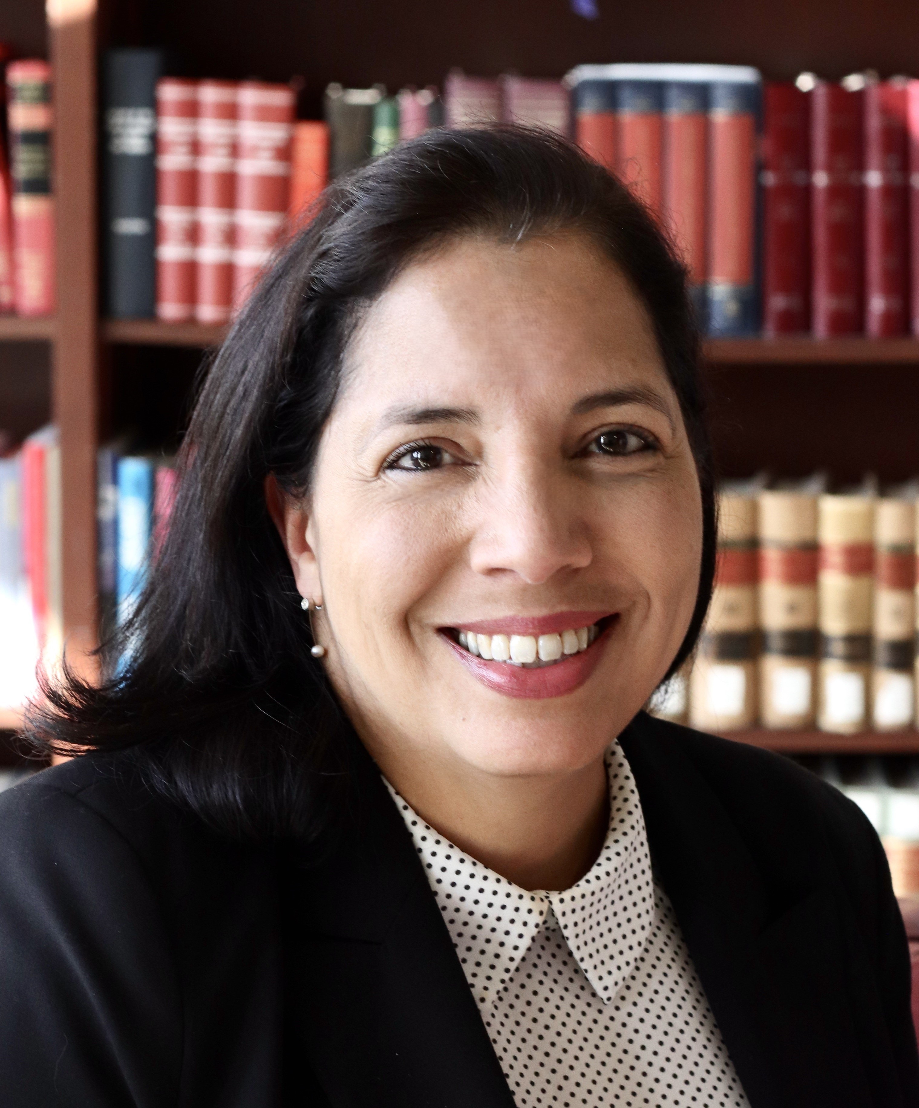 Professional headshot of a woman with shoulder length black hair wearing a dark blazer and a light polka-dot button-up shirt, seated in front of bookshelves filled with legal books. The woman is facing the camera and smiling, and the image is lit with soft, natural-looking light.